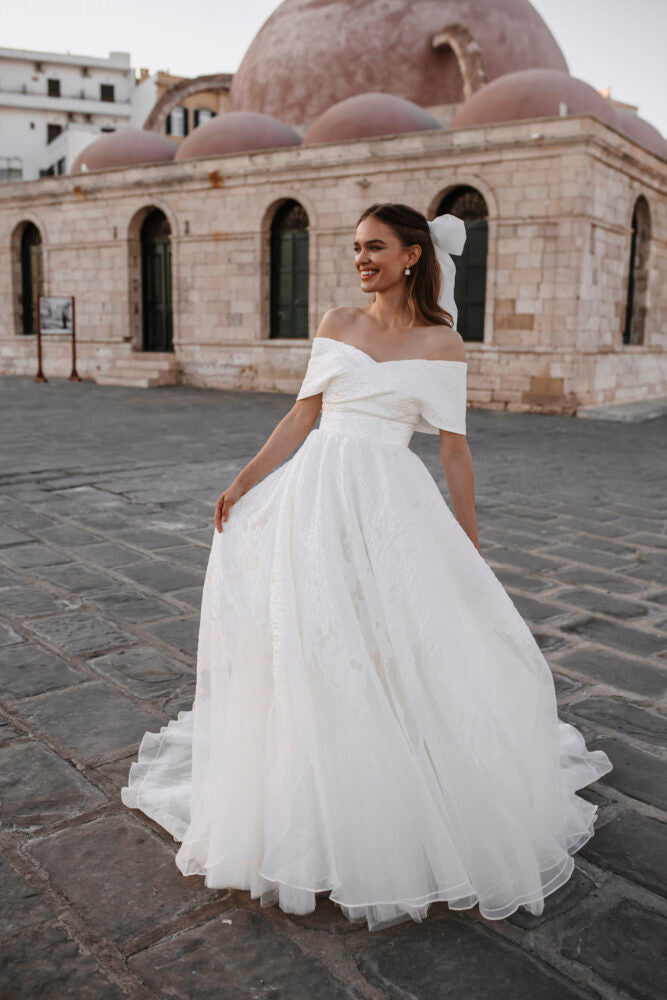 Feminine off-the-shoulder ball gown in ivory colour  with a heart-shaped neckline. Features a structured fabric, draped bodice, an open back and a full skirt with a long train.